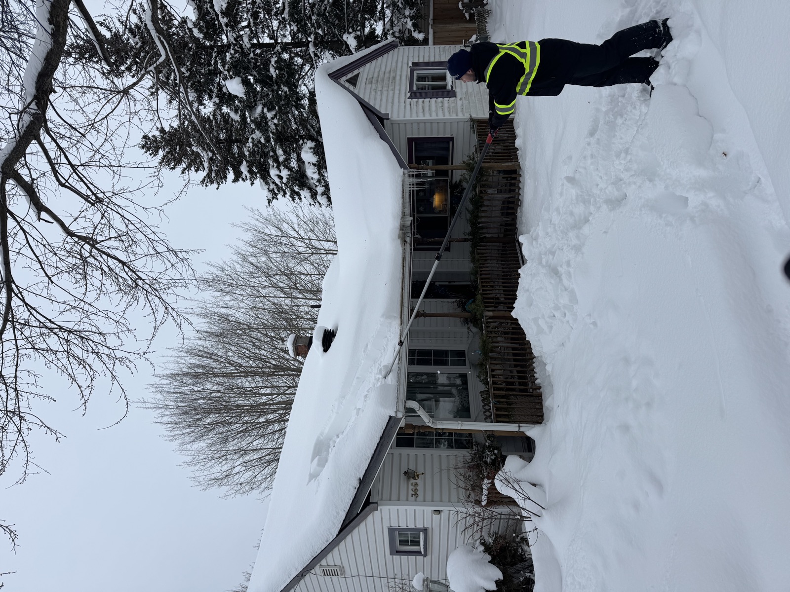 Crew member raking heavy snow off a bungalow roof with icicles along the eaves