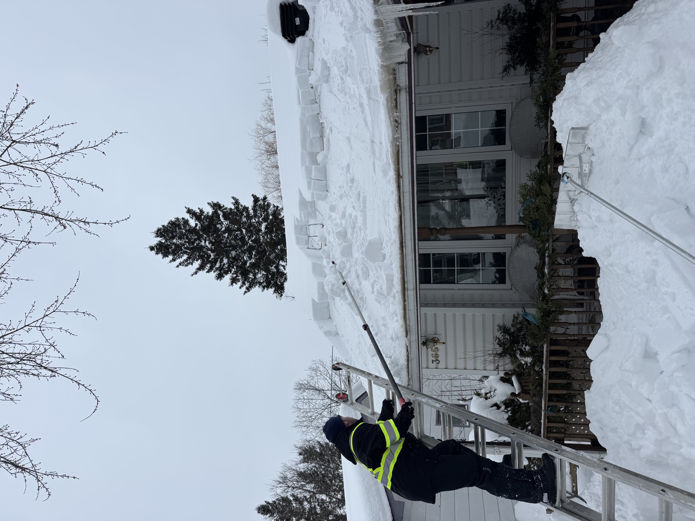 Crew member clearing snow from a roof edge with a roof rake from a ladder